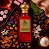 A luxurious red glass perfume bottle with a gold crown-shaped cap, labeled “Royale Noir – Candy Shop Extrait de Parfum, 60 mL – 2.2 FL. OZ.” The bottle is arranged on a rich burgundy background surrounded by candy and gourmand ingredients: assorted colorful jelly beans, marshmallows, pink chewing gum, caramel candies, pieces of brown sugar, vanilla beans, cacao beans, white jasmine flowers, wood sticks, a small bowl of white powder, dark dried fruits, green leaves, and a purple violet flower.