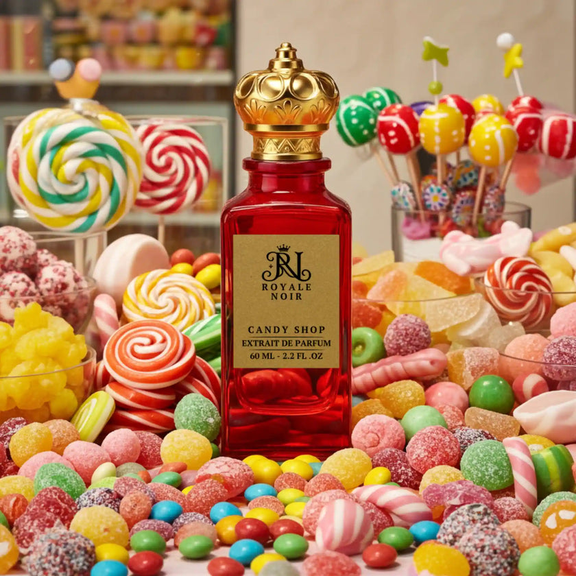 A luxurious red glass perfume bottle with a gold crown-shaped cap, labeled “Royale Noir – Candy Shop Extrait de Parfum, 60 mL – 2.2 FL. OZ.” The bottle is arranged on a rich burgundy background surrounded by candy and gourmand ingredients: assorted colorful jelly beans, marshmallows, pink chewing gum, caramel candies, pieces of brown sugar, vanilla beans, cacao beans, white jasmine flowers, wood sticks, a small bowl of white powder, dark dried fruits, green leaves, and a purple violet flower.